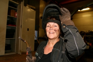 Woman with welding torch and gloves, welding mask tilted up, smiling at camera